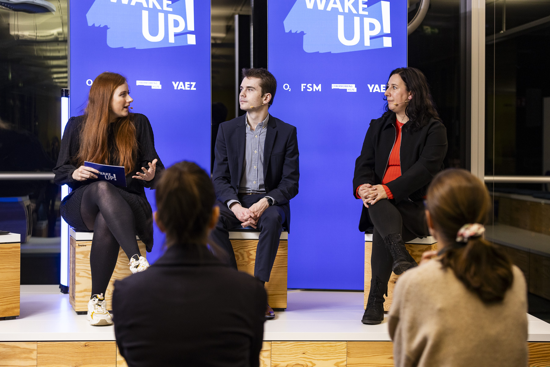 Moderatorin sitzt mit zwei Referenten auf der Bühne und stellt ihnen eine Frage.