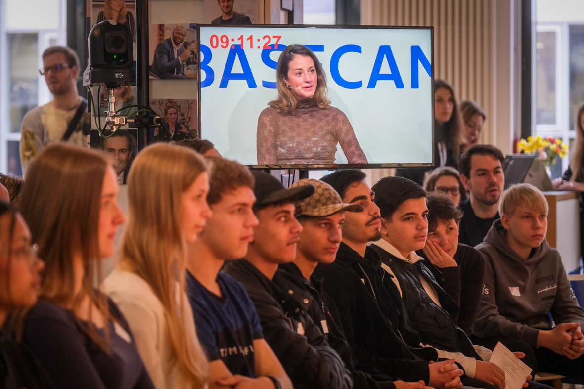 Schüler:innen sitzen im Publikum, auf einem Bildschirm sieht man Politikerin